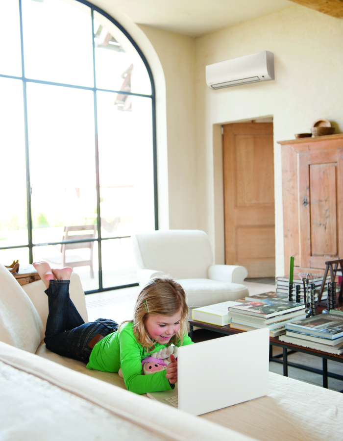 Klimaanlagen Wandgerät an hellgelber Wand - kleines Mädchen in grünem Pullover liegt auf Sofa und guckt auf Laptop